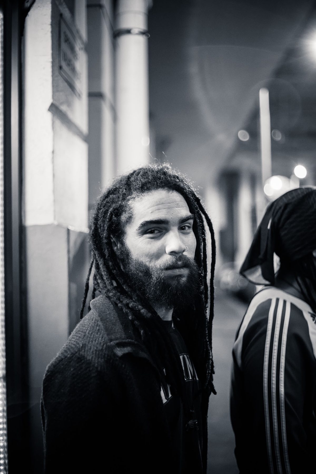 A man with dreadlocks and a beard stands on a street corner at night