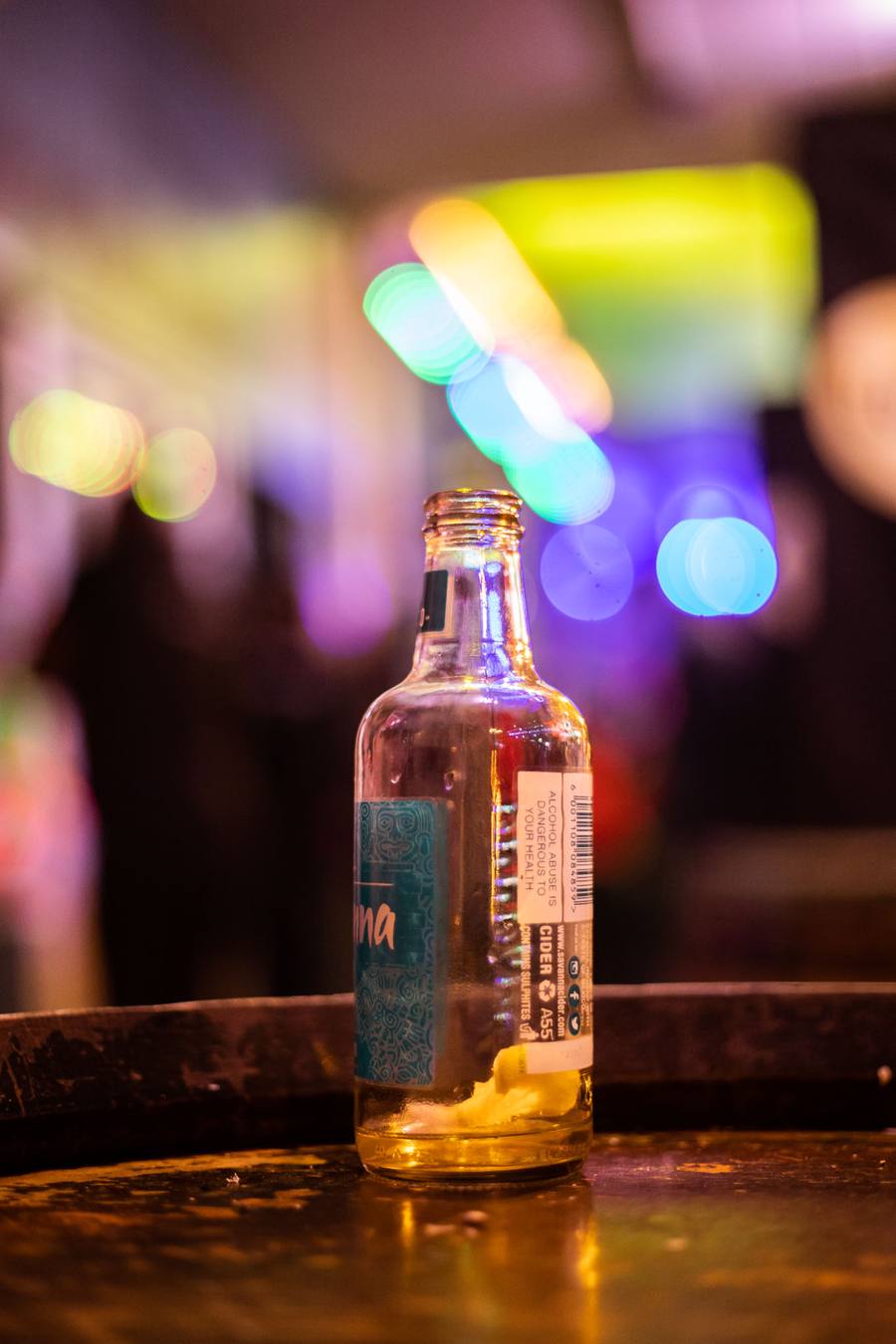 An empty bottle of cider on a wooden table with blurred lights in the background