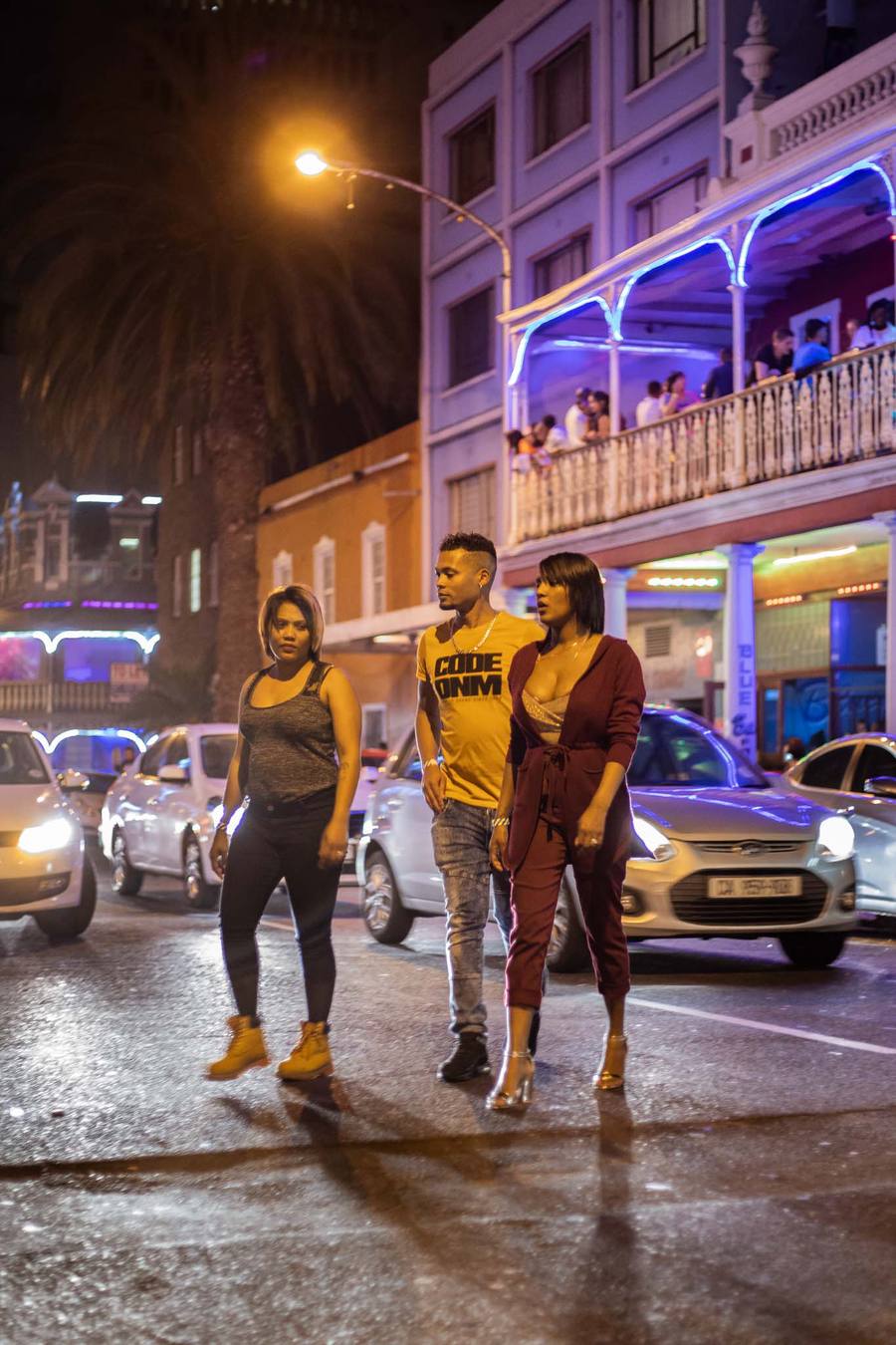 Three people walk along a busy street at night, with cars and buildings in the background