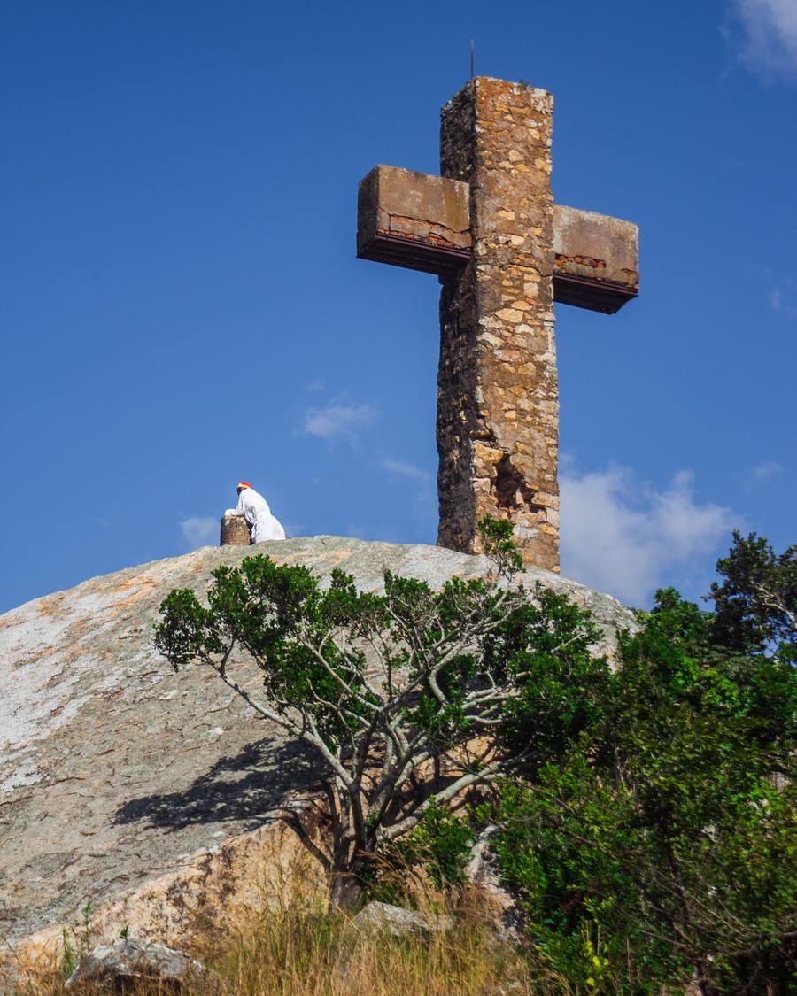 A large stone cross on a hill with a person in white clothing kneeling in the foreground