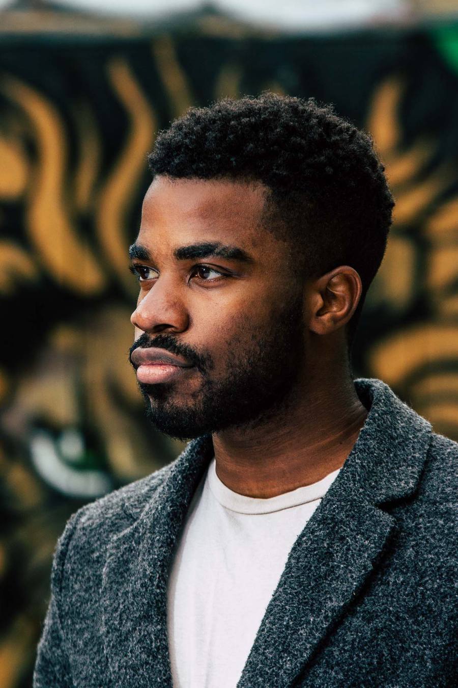 Man with beard and moustache wearing a white t-shirt and grey jacket, standing in front of a tiger mural.