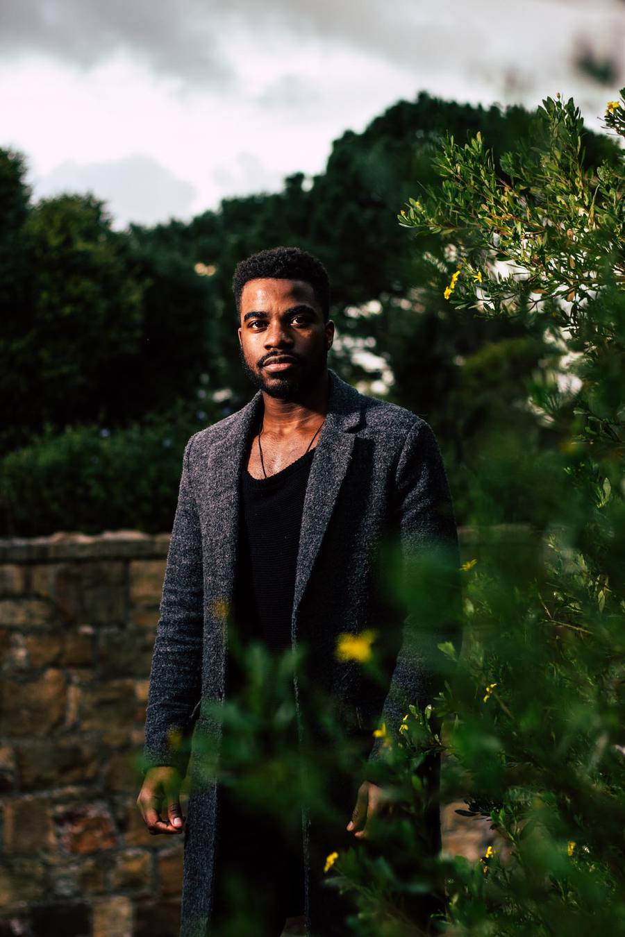 Man in grey coat and black top stands in front of stone wall and greenery