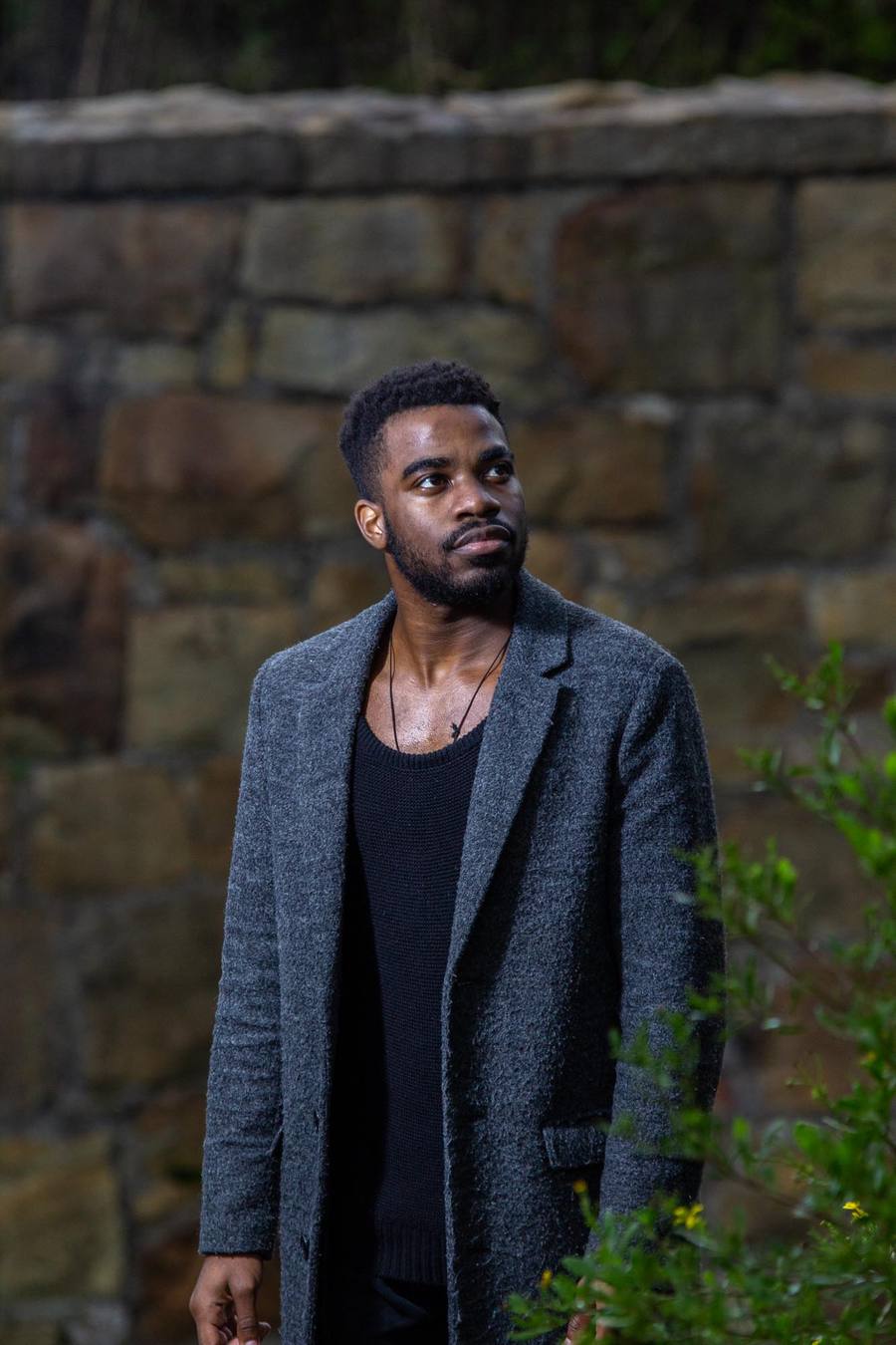 Man in black top and grey coat standing in front of stone wall