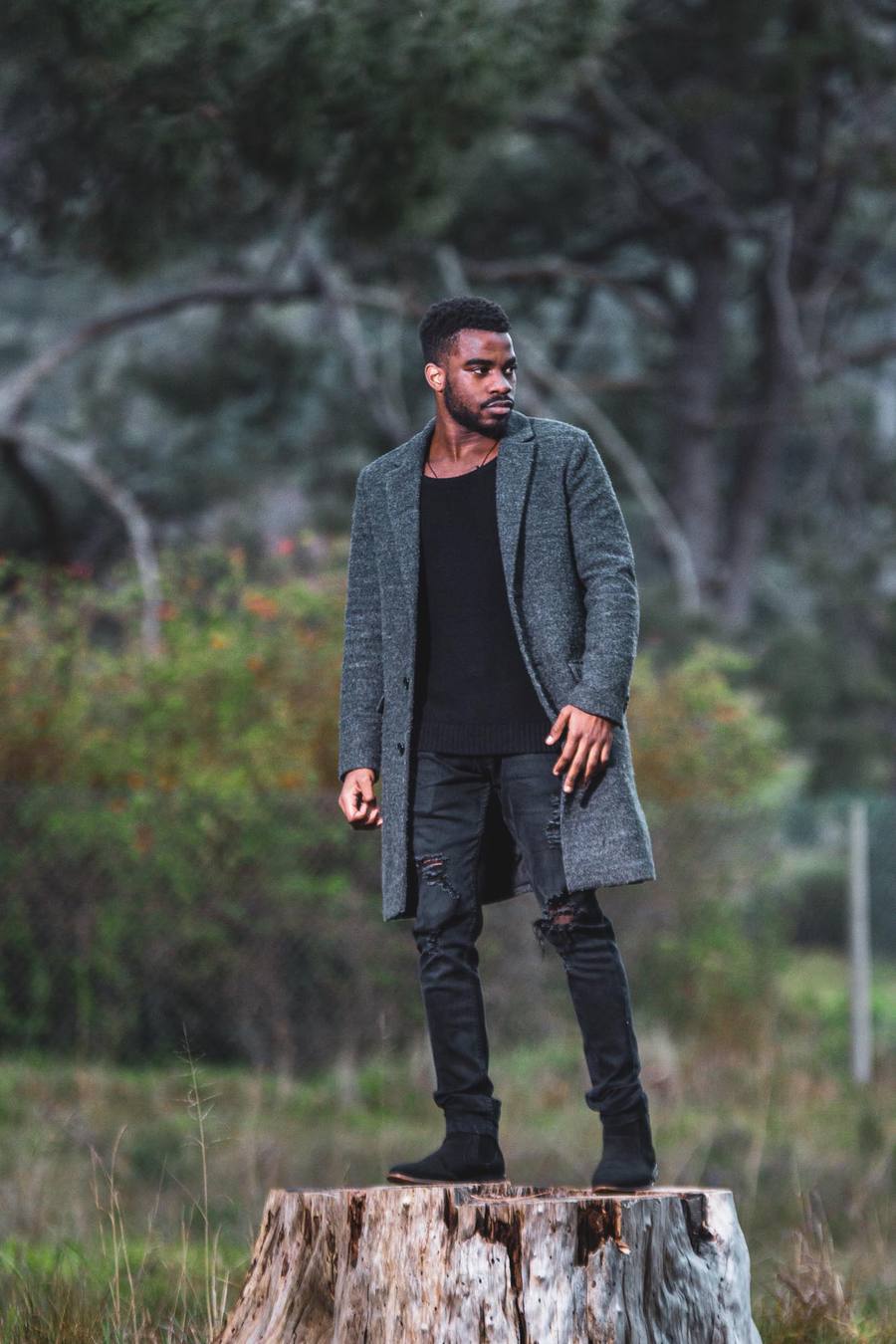 Man in grey coat and black top stands on tree stump in woodland