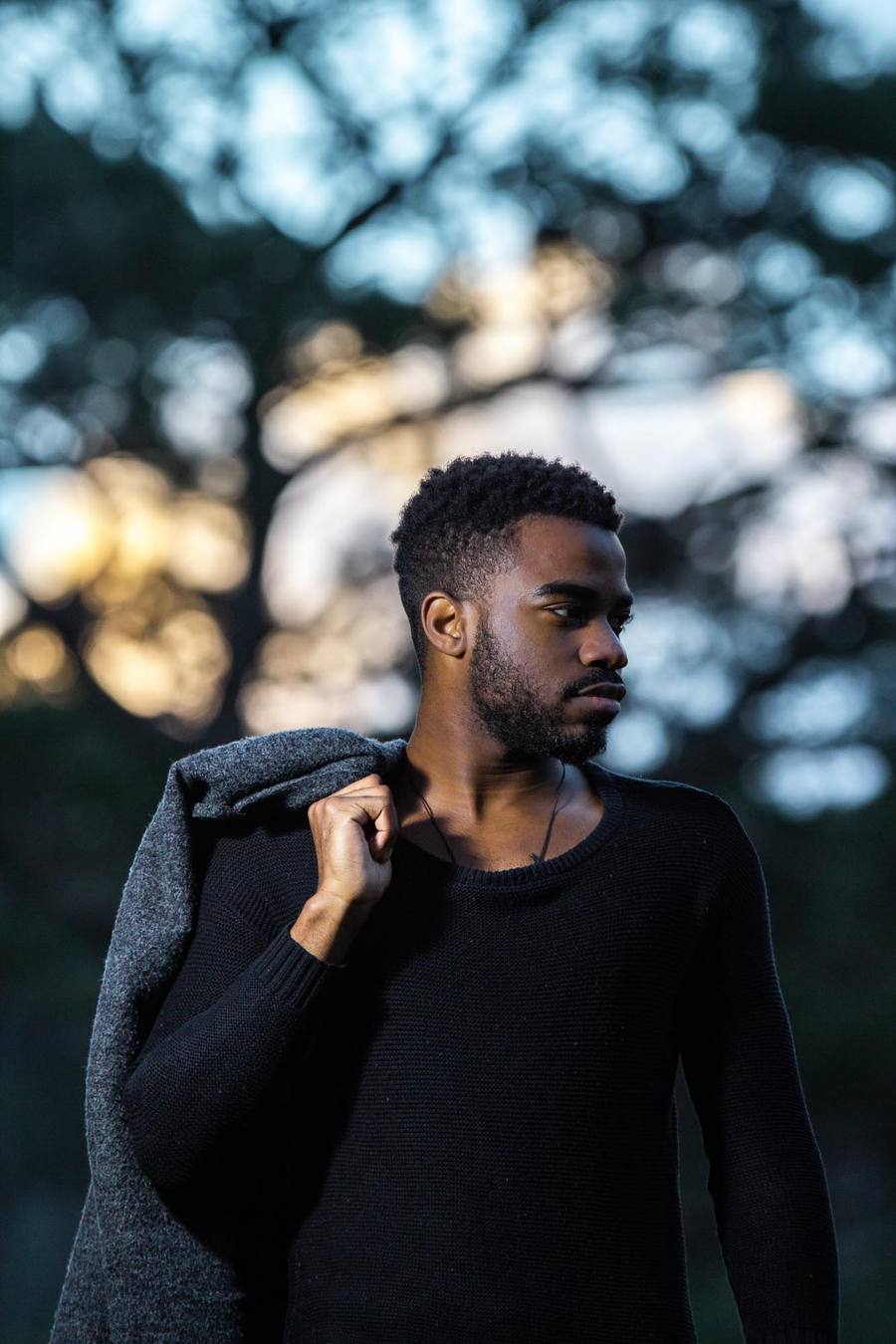 Man in black jumper and grey jacket standing outside with trees in background