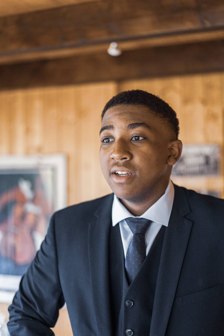 A man in a dark suit and tie smiles at the camera in a room with wooden walls.
