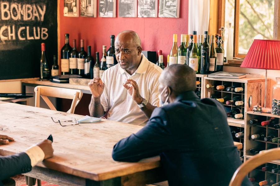 Three men sit around a table in a room with a chalkboard and wine bottles.