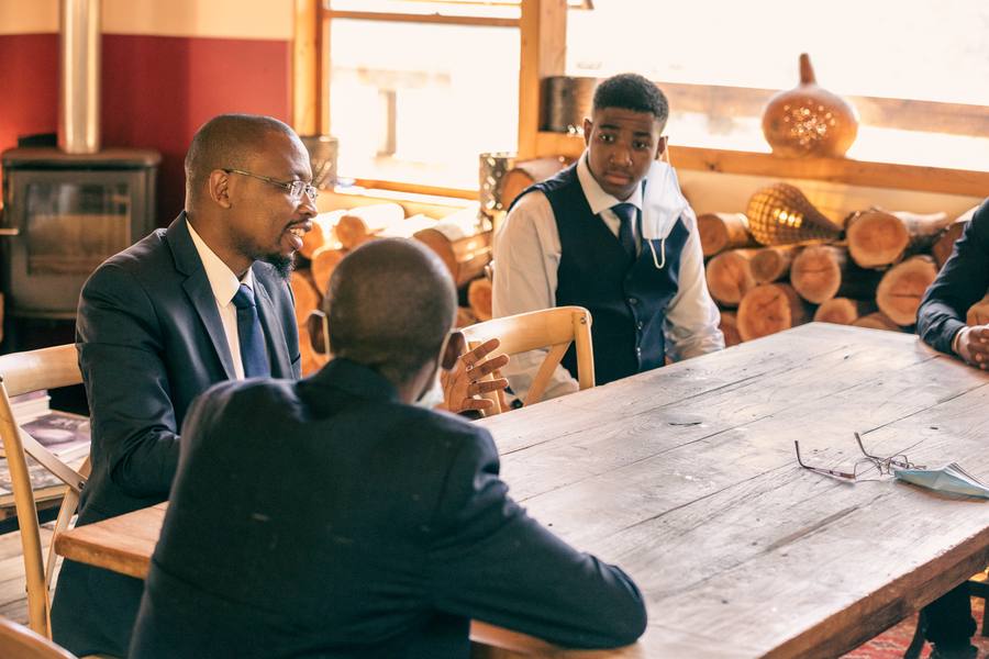 Three men in suits sit around a wooden table, one wearing a mask
