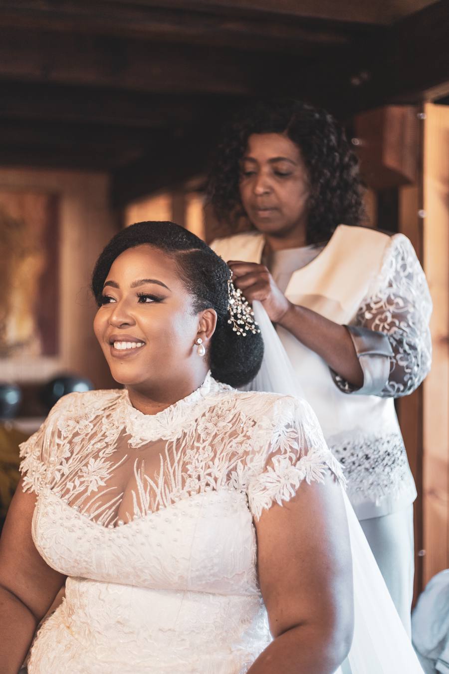 Bride smiles as woman adjusts veil behind her in a room with wooden walls.