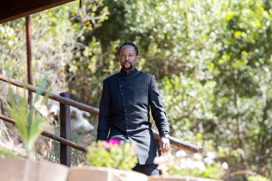 Man in black suit standing on wooden railings with flowers and greenery in background