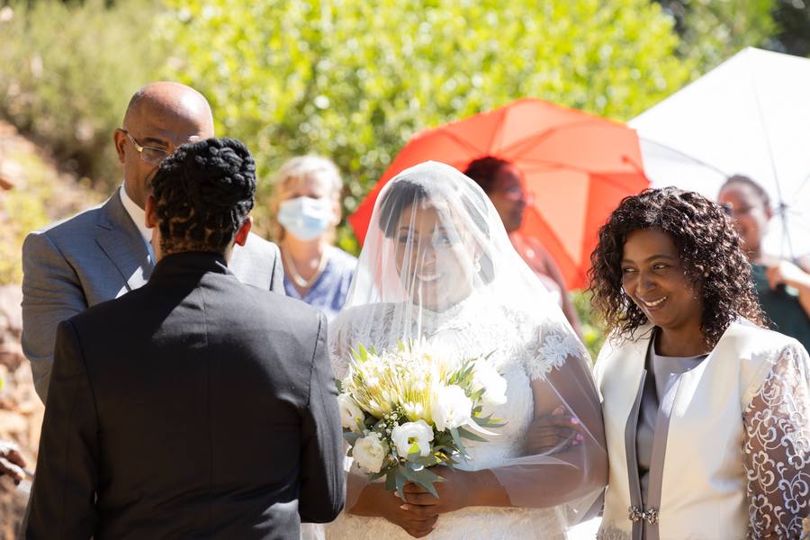 Bride smiles at groom with bouquet, veil, and guests in background.
