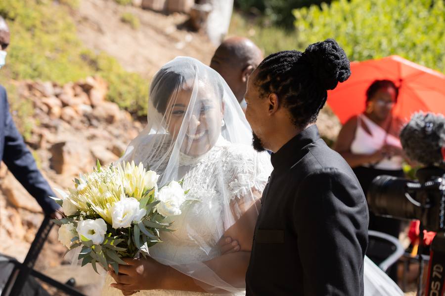 Bride and groom share a moment, surrounded by guests and greenery.