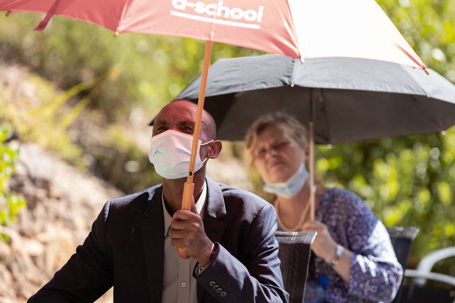 A man and woman wearing masks sit under umbrellas in a garden.