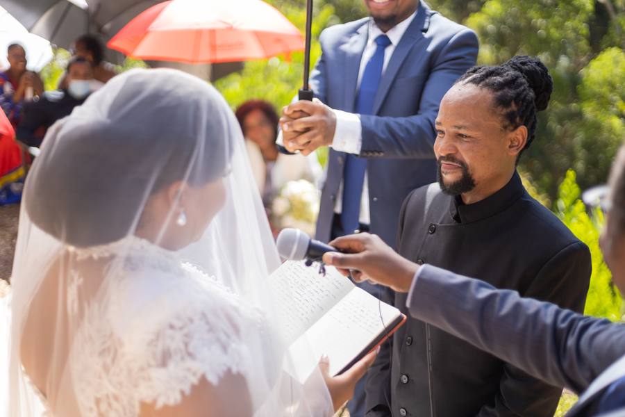 A bride and groom exchange vows while guests look on, one holding an umbrella.