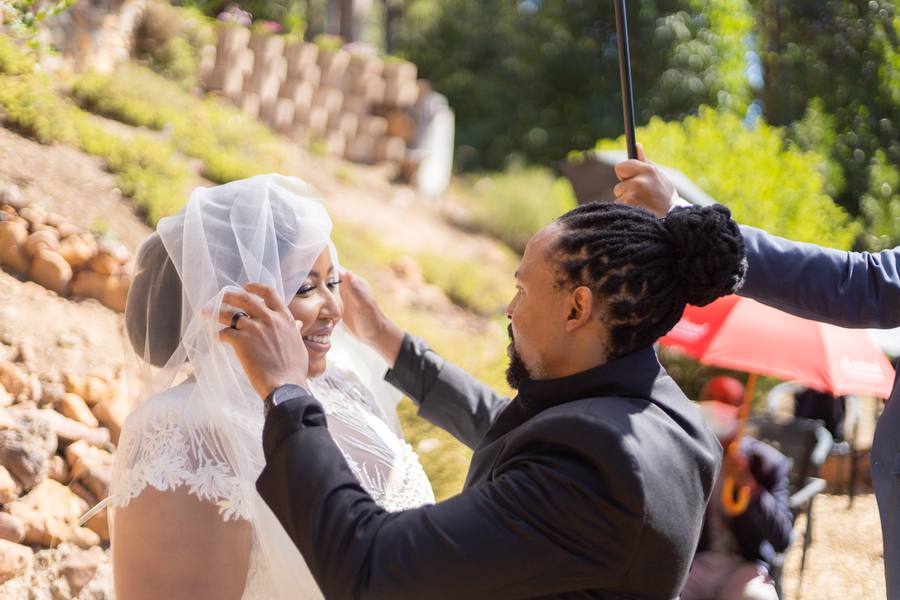 A groom adjusts his bride's veil while holding an umbrella over them.