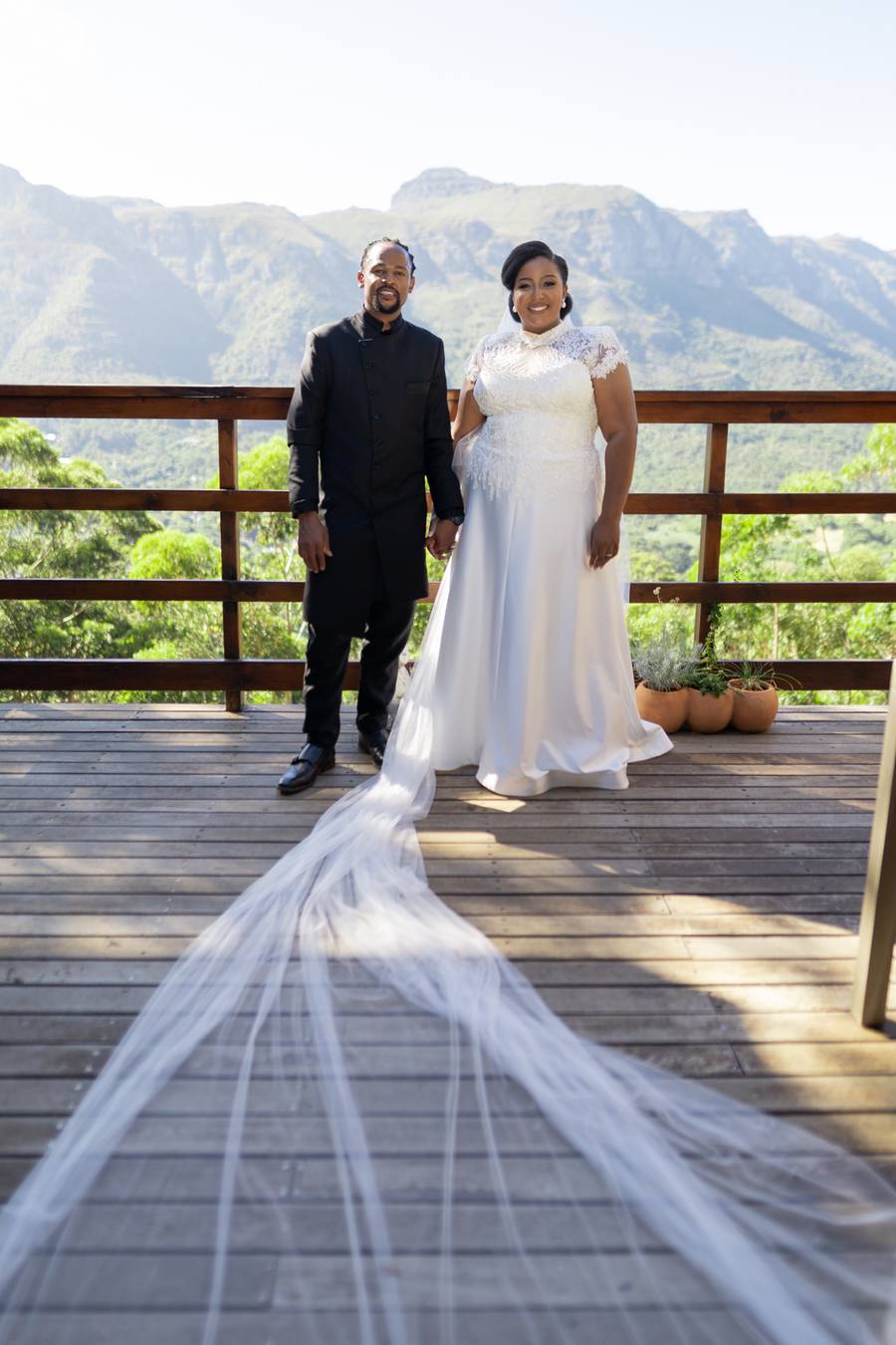 A bride and groom stand on a wooden deck with mountains in the background.