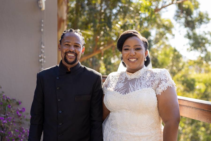 A smiling couple stands on a balcony with trees in the background