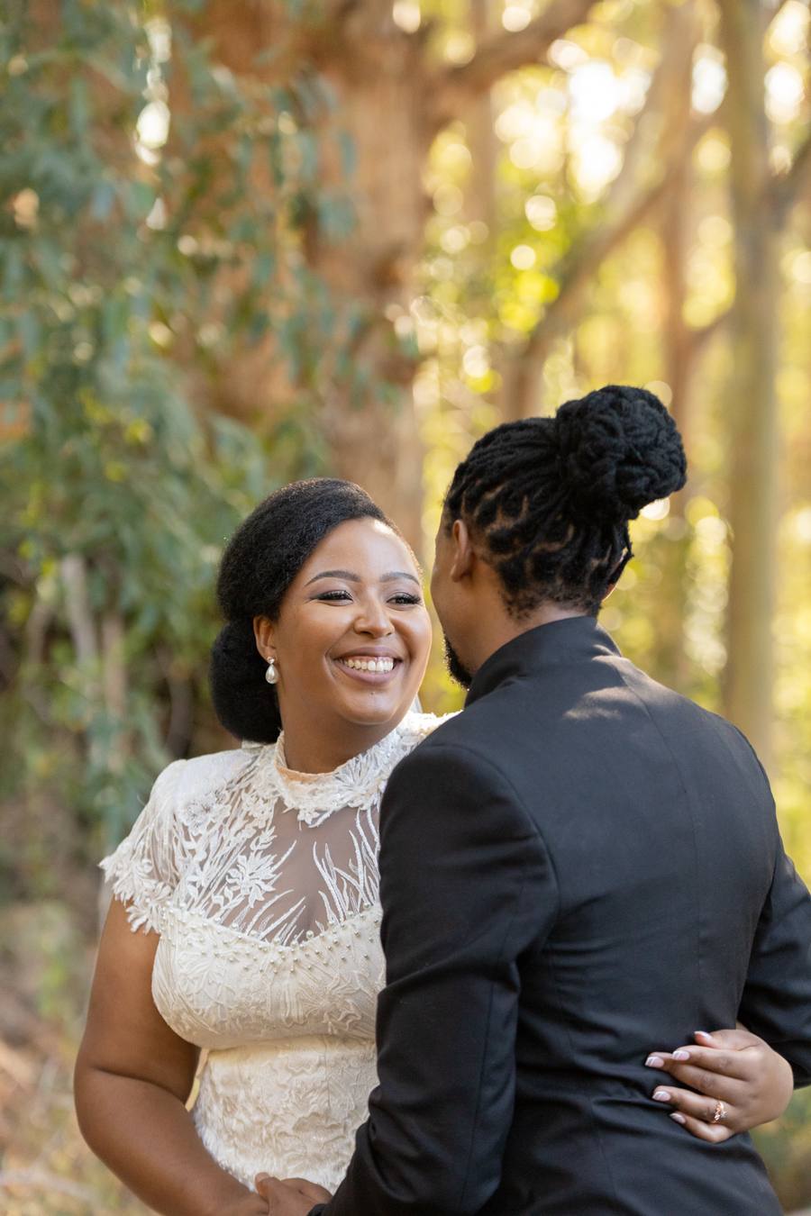A smiling bride and groom stand close together in a forest.