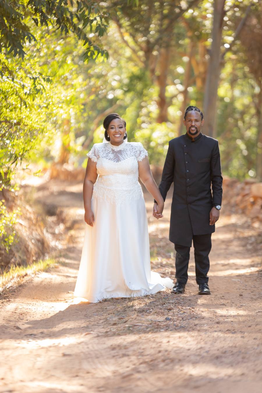 A bride and groom hold hands on a dirt path in a forest.