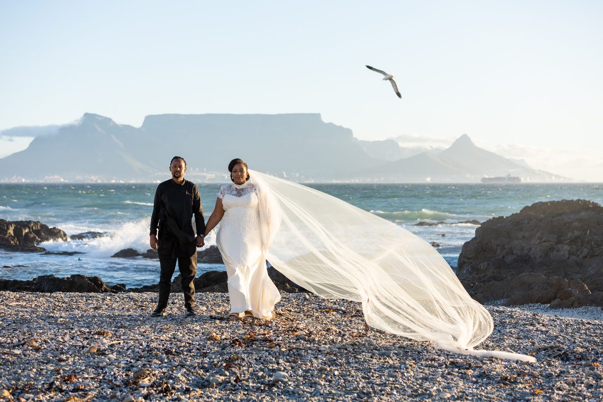A bride and groom stand on a pebble beach with mountains in the background