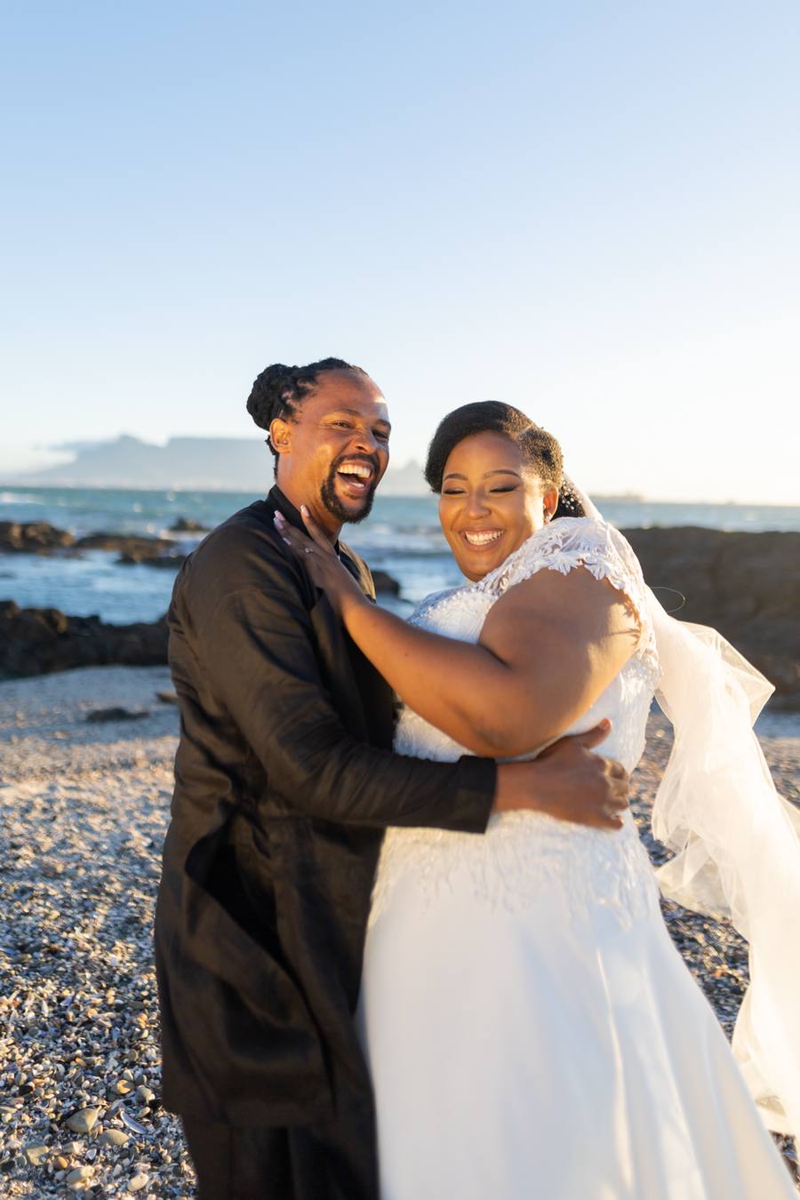 A newlywed couple embrace on a pebble beach with a city skyline in the distance.