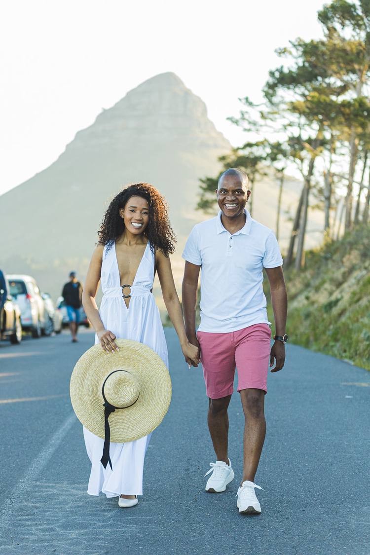 A couple holding hands and smiling, walking on a road with a mountain in the background.