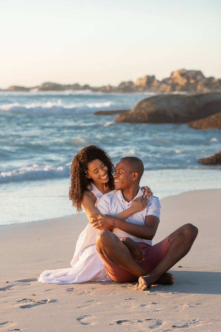 Couple sitting on beach, smiling at each other, ocean in background