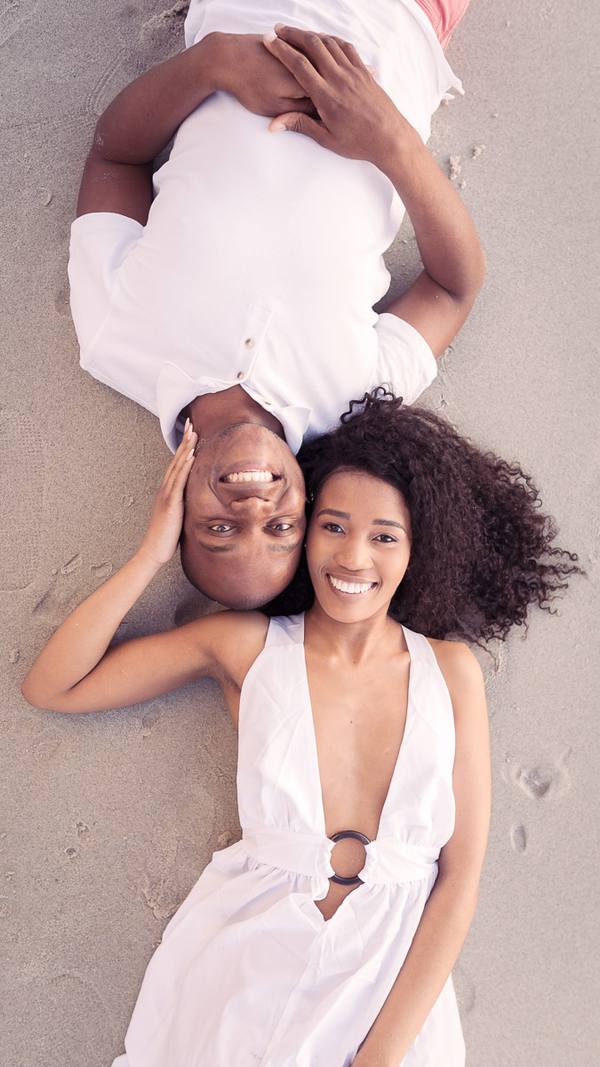 A man and woman lying on a beach, both smiling and wearing white clothes