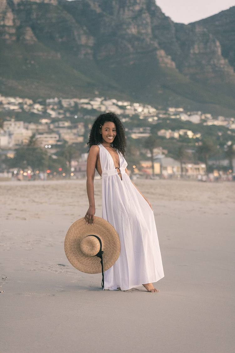 Woman in white dress holding a straw hat on a beach with mountains in the background