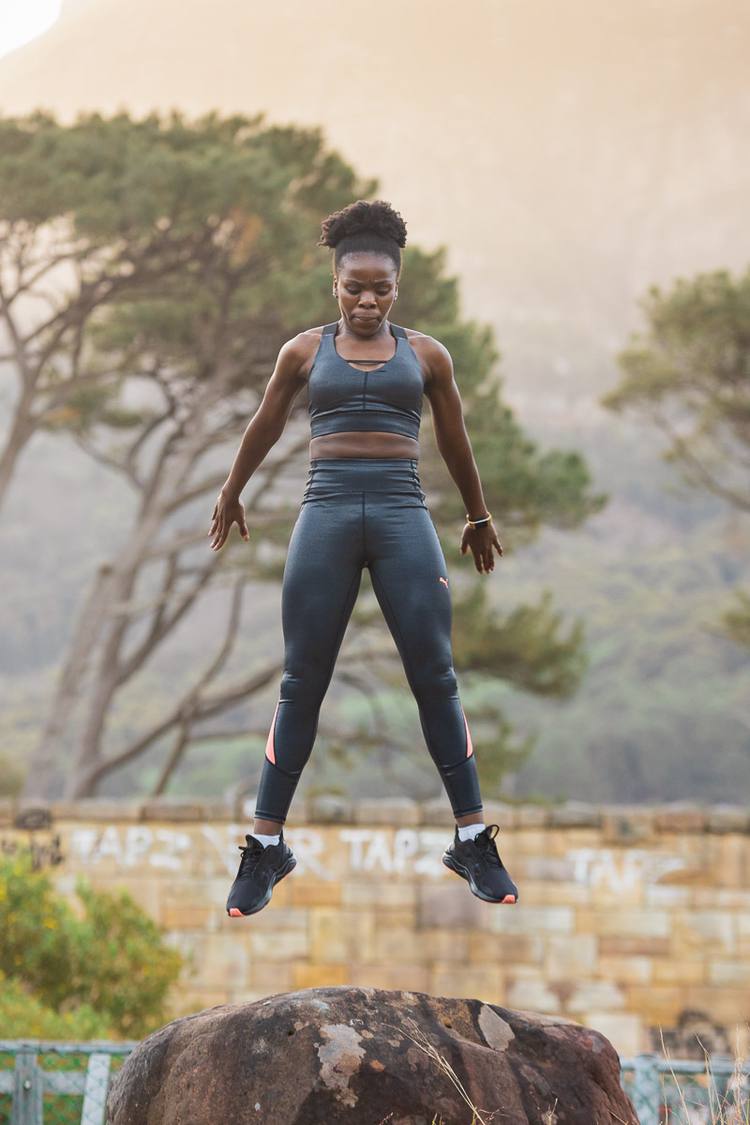Woman in athletic wear jumping over rock with trees and wall in background