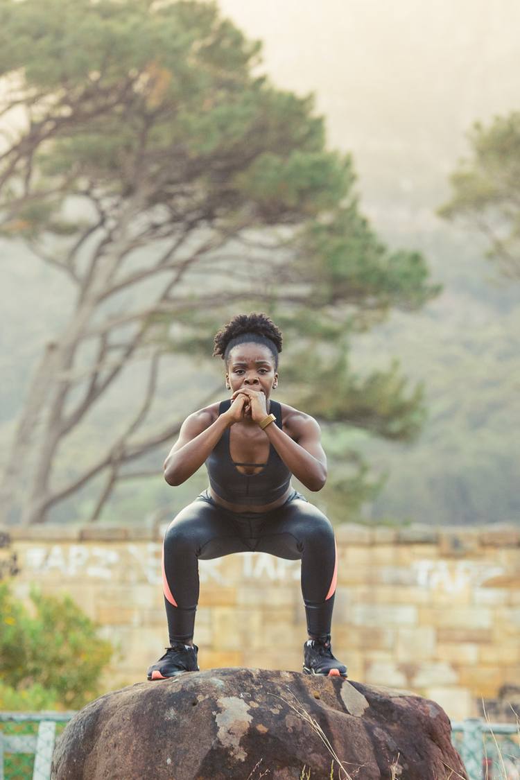 Woman in black and pink leggings squatting on a rock with trees in the background