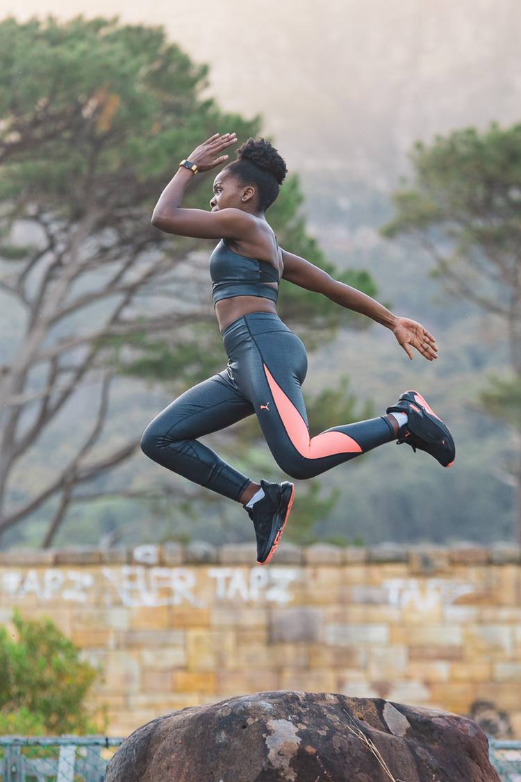 Woman in sports gear jumping over rock with graffiti wall in background.