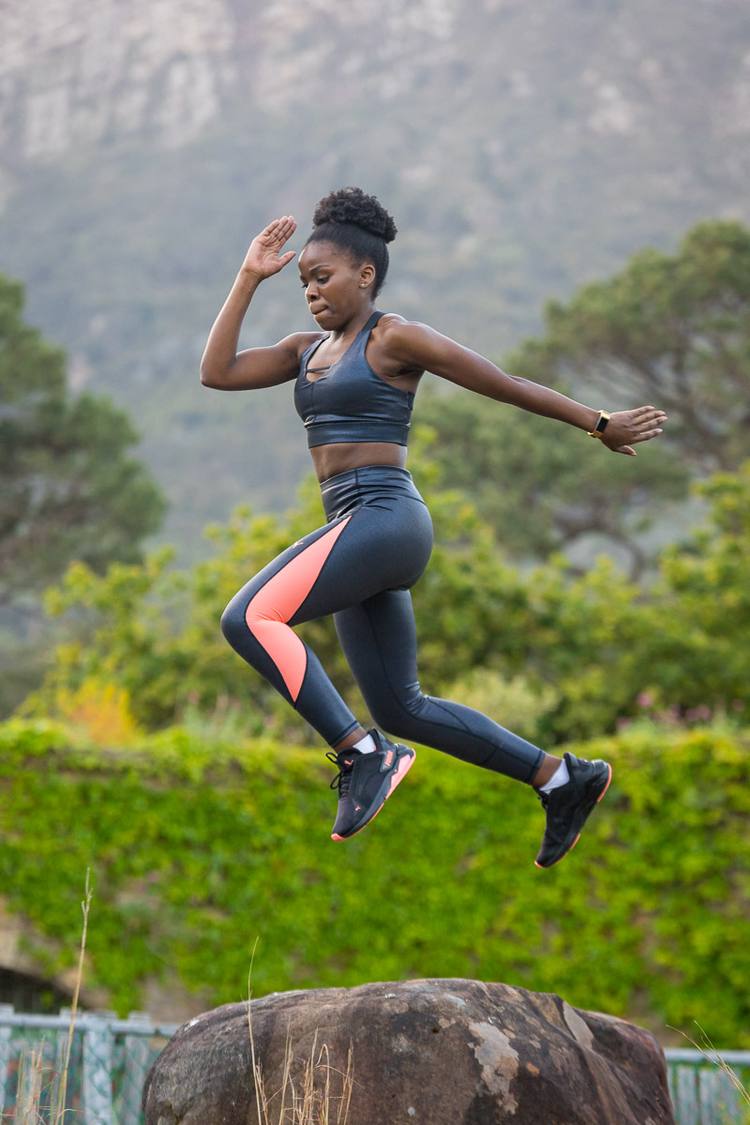 Woman jumping over rock in outdoor setting with greenery and mountains in background