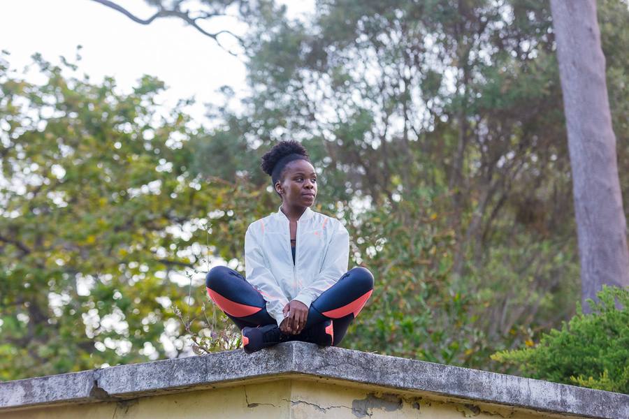 Woman in white jacket and pink leggings sitting cross-legged on concrete wall, trees in background
