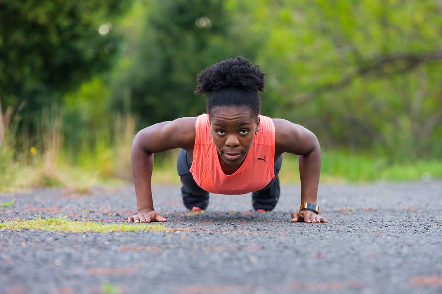 A woman in an orange vest performs push-ups on a gravel path with trees in the background.