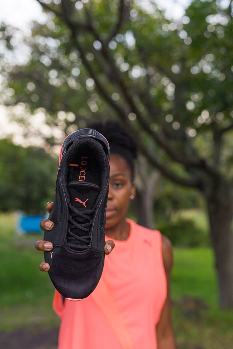 Woman in pink top holds up black Puma sneaker with orange accents