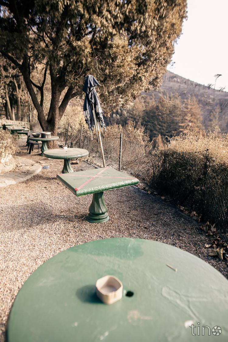 Green picnic tables and a closed umbrella in a park with a burnt hillside in the background