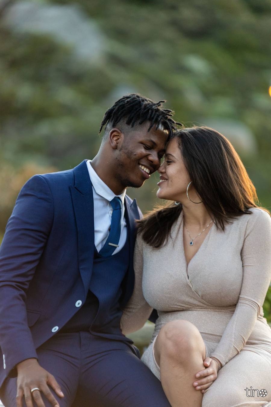 A man and woman sit close together, smiling at each other, with a natural background.
