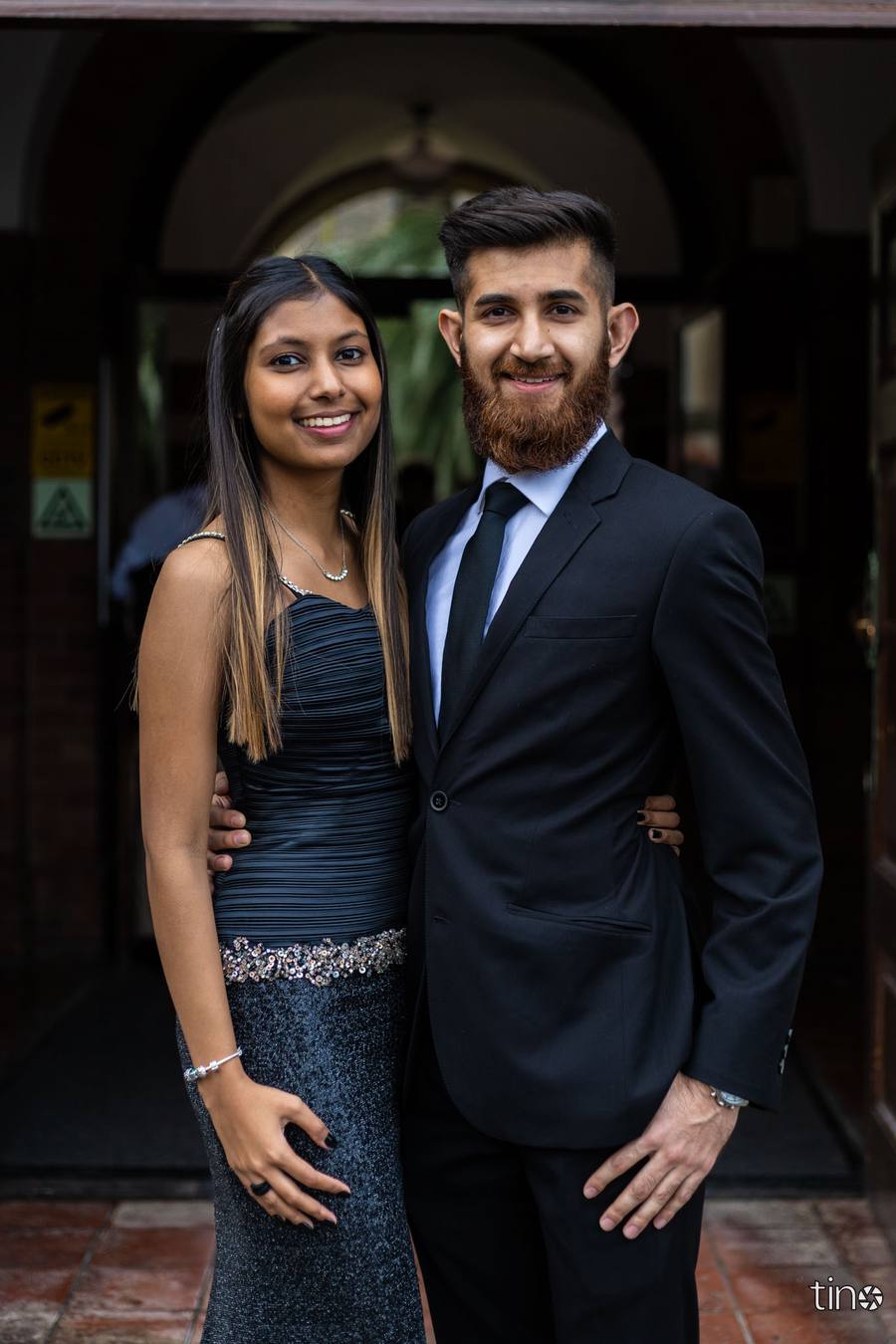 A man and woman stand close together, smiling, in front of a building.
