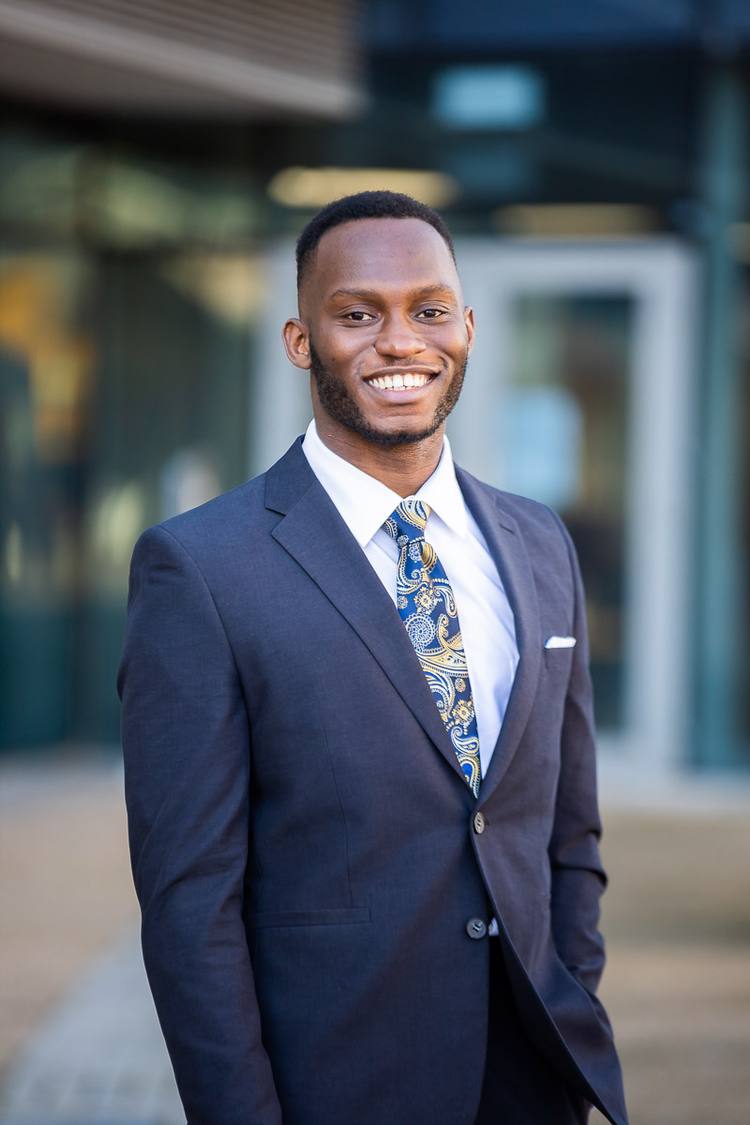A man in a navy suit and paisley tie stands outside a building, smiling.