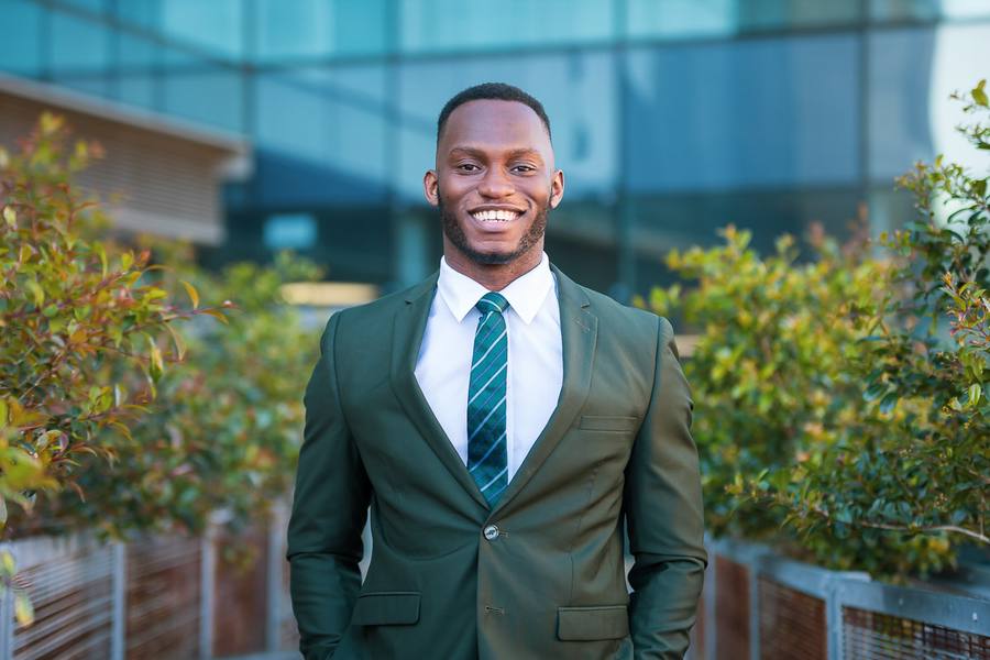 Man in green suit and striped tie smiling outside with plants and building in background