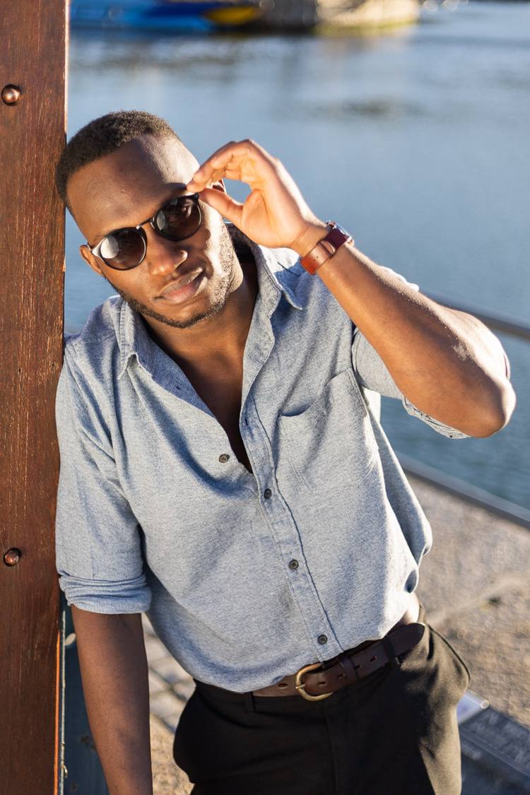Man in sunglasses and grey shirt leaning against wooden post by water