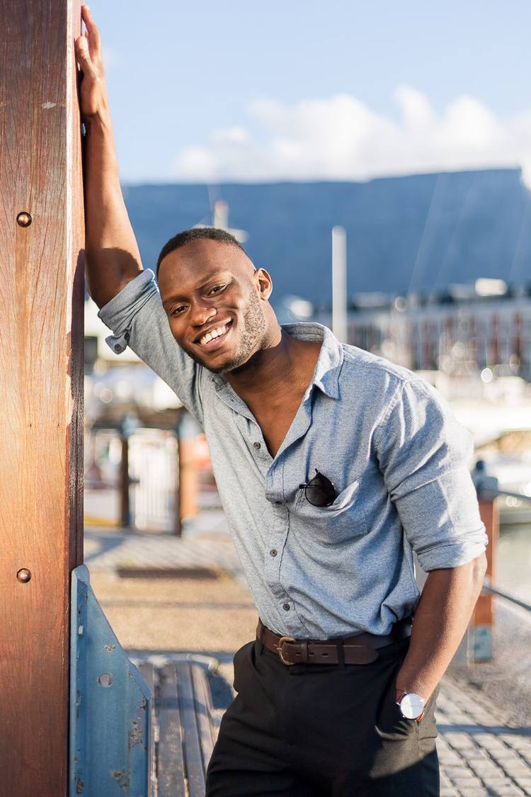A man in a grey shirt and brown belt smiles at the camera on a dock.