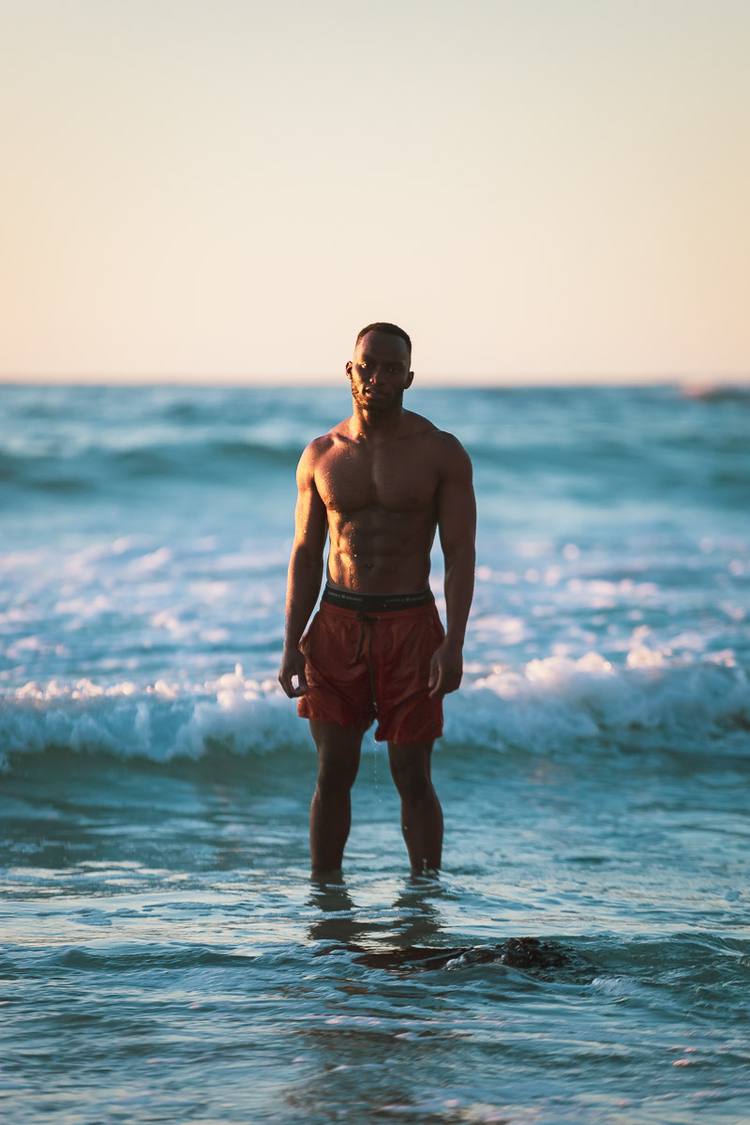 A man stands in the ocean at sunset, wearing red shorts and no shirt.