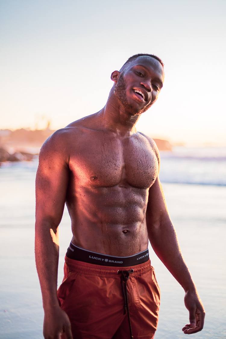 A man stands on a beach, wearing orange shorts and a black waistband, smiling at the camera.