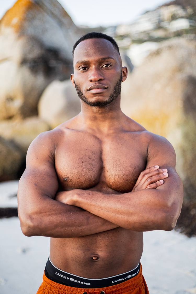 A man with muscular arms and torso stands on a beach with his arms crossed