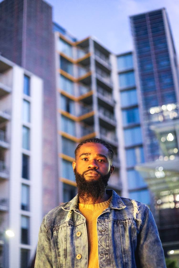 Man with beard wearing yellow jumper and denim jacket standing in front of modern buildings