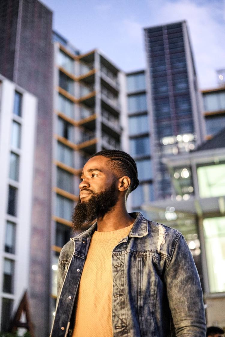 Man with beard and denim jacket stands in front of modern buildings