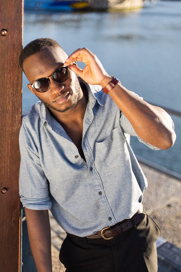 Man in sunglasses and grey shirt leaning against wooden post by water