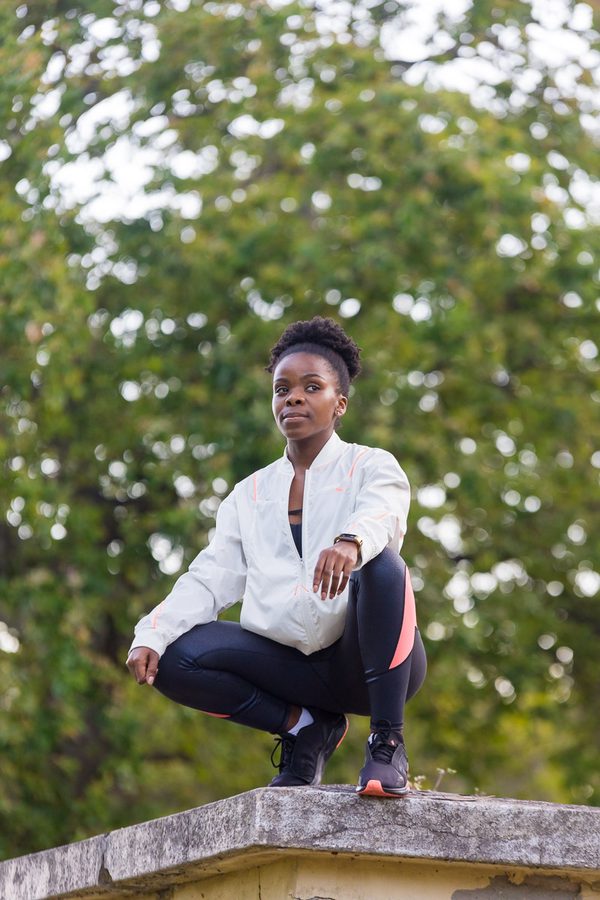 Woman in white jacket and black leggings crouching on concrete wall with trees in background